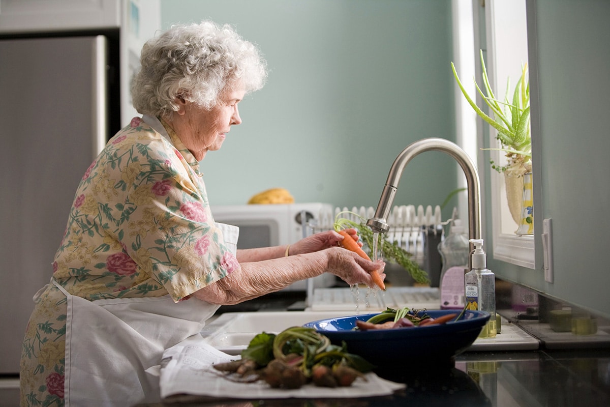 a senior woman in senior living facility wearing an apron washing vegestables 